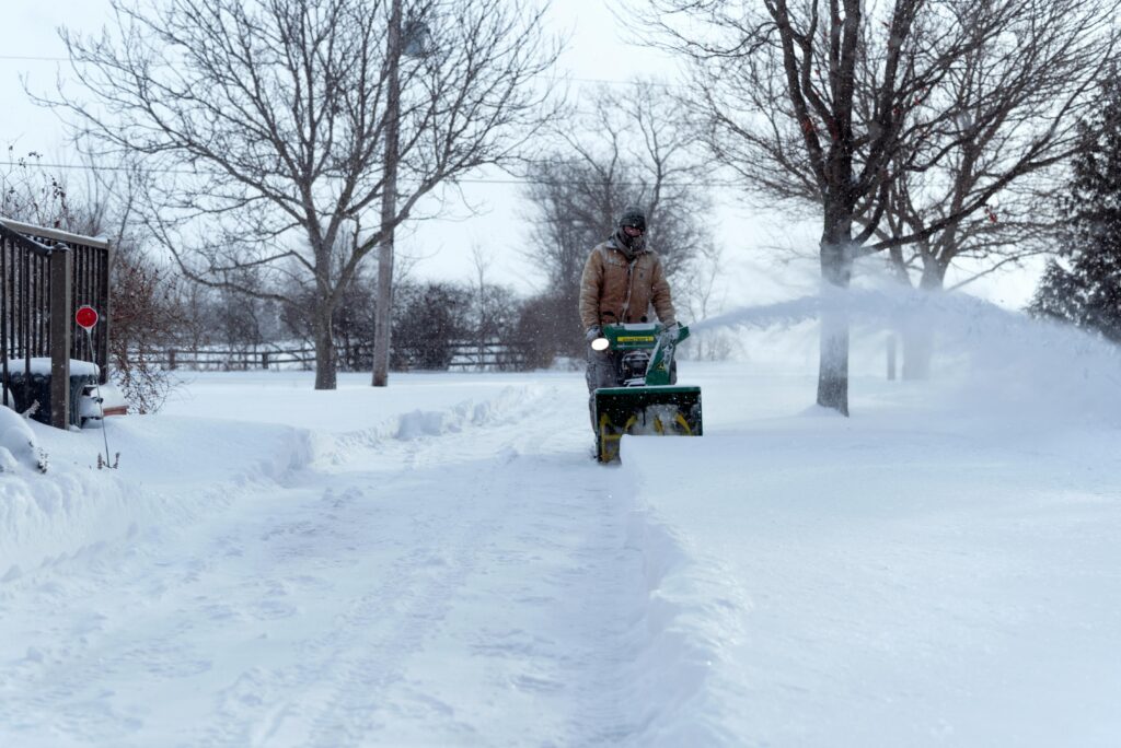 A person operates a snowblower on a snow-covered driveway in Essex, Ontario, during winter.